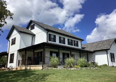 Two-story white house with black shutters and a porch, surrounded by green grass and plants under a blue sky with fluffy clouds.