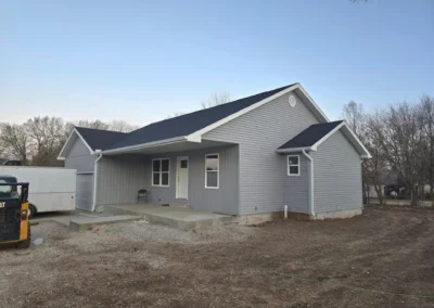 A newly constructed gray house with a dark roof, front porch, and gravel yard, surrounded by sparse trees in the background.